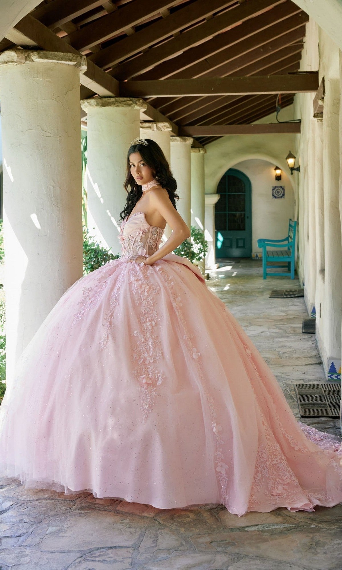 A woman stands on a stone porch wearing the Quinceanera Dress JT1497J by Julietโa voluminous pastel-pink ballgown adorned with delicate floral lace appliquesโposing with one hand on her hip and looking back over her shoulder.